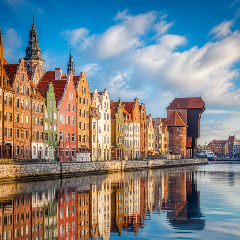 AI-generated illustration of a Gdansk-inspired historic waterfront with tall slender colourful merchant houses and a medieval timber harbour crane reflected in calm blue water